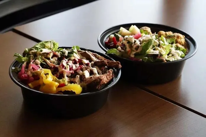 Two Indian rice bowls with grilled meat, greens, pickled onion, and drizzled sauces on a wooden table at The Indian Belly in Olathe