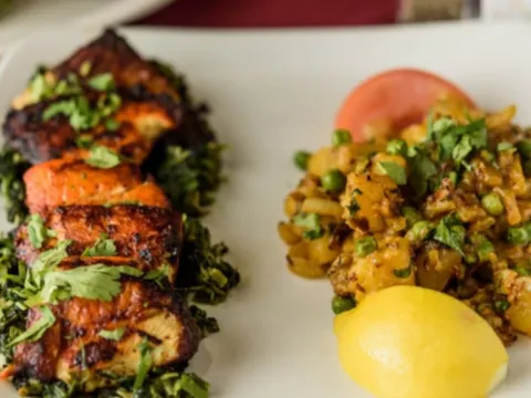 A white plate with charred tandoori chicken over sauteed greens beside a spiced potato-and-pea side and lemon at Silver Coin Indian Grill in Birmingham