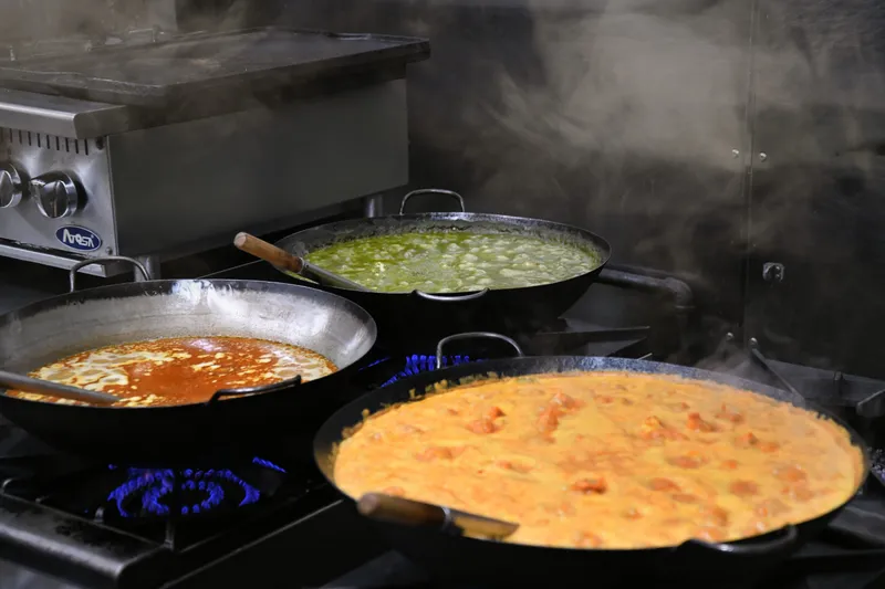 Three steaming karahi pans of curry — green saag, a red masala and an orange butter-chicken-style gravy — cooking on the stove in the kitchen at Masala in Missoula