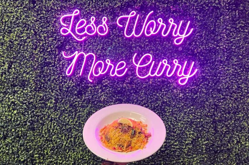 Plate of biryani held up in front of the pink "Less Worry More Curry" neon sign on a greenery wall at The Curry Modern Indian in Huntsville